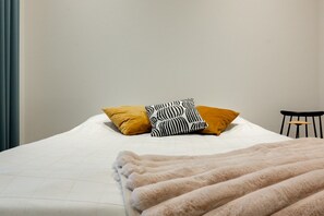 Un lit avec un matelas blanc, deux oreillers, une couverture beige moelleuse et une chaise noire.