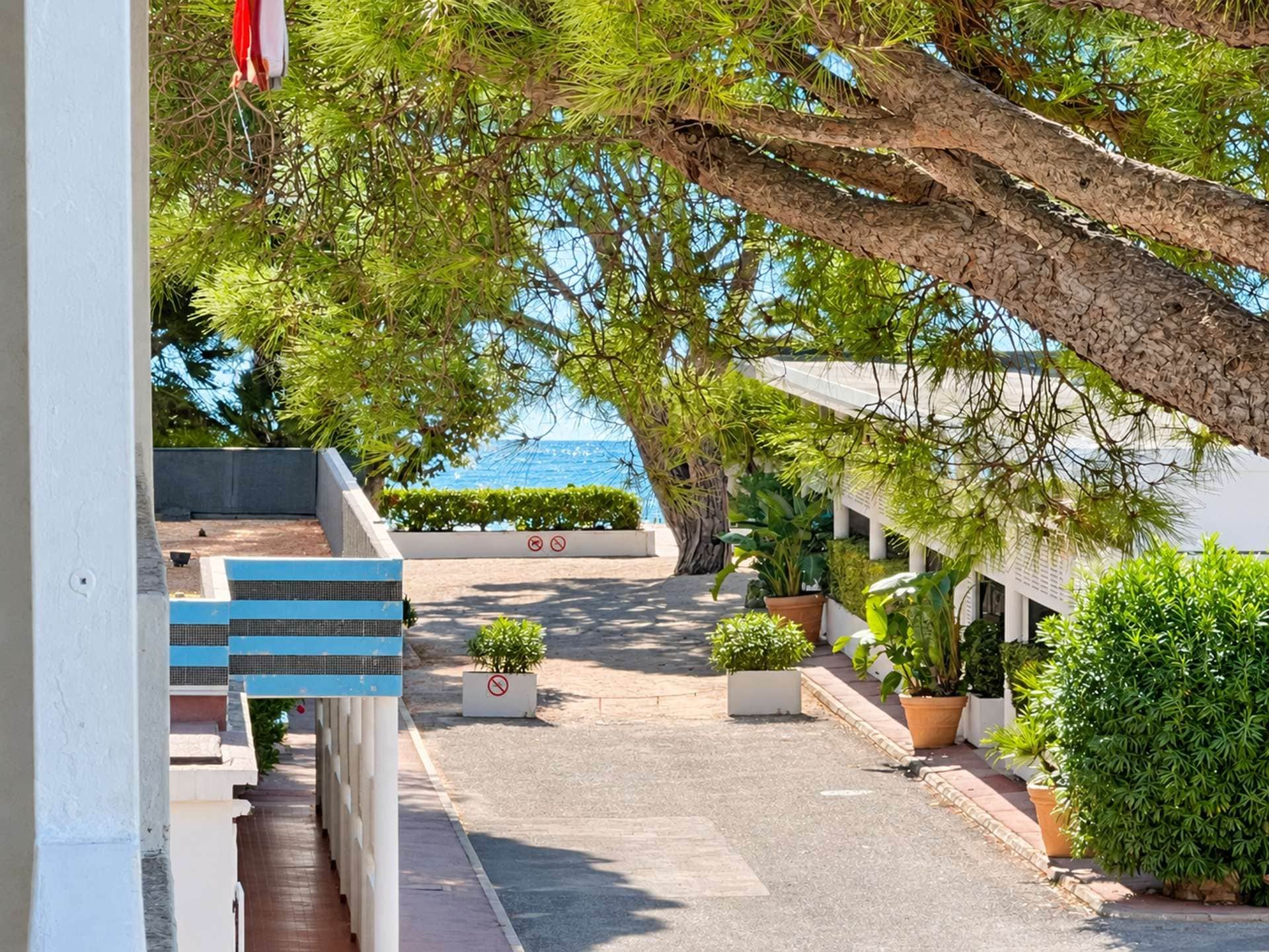 A pathway leading to a building with a view of the sea. There are potted plants along the path and a staircase on the left.