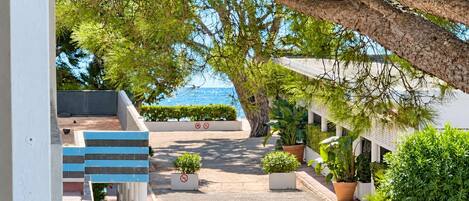 A pathway leading to a building with a view of the sea. There are potted plants along the path and a staircase on the left.