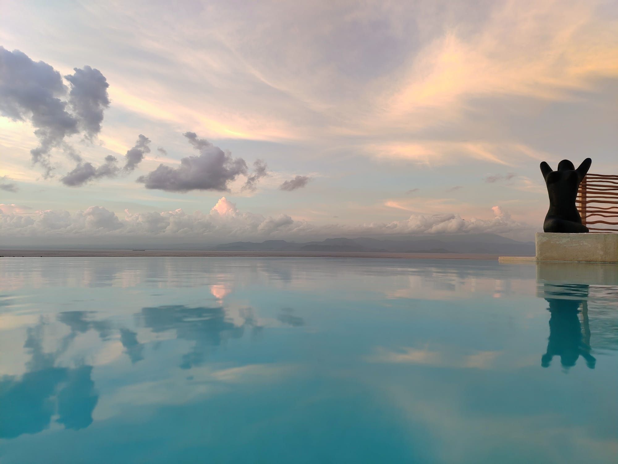 Una piscina a sfioro tranquilla che riflette il cielo e le nuvole, con un elemento scultoreo sulla destra.
