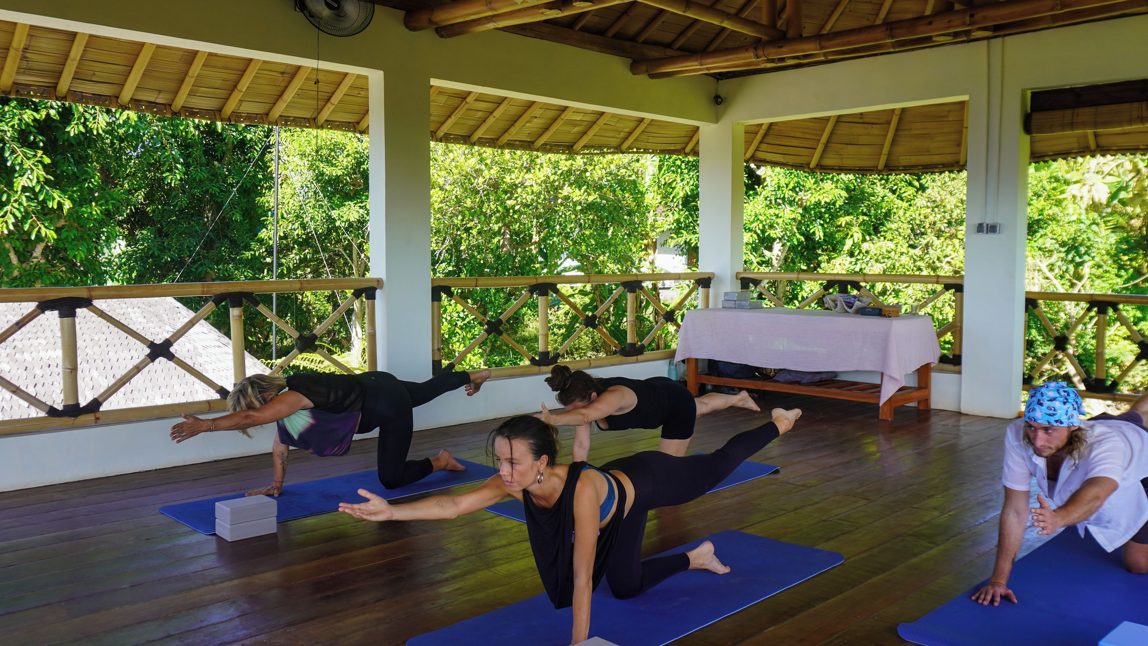 Un gruppo di persone pratica yoga su tappetini in un'area esterna coperta, con pavimento in legno e un tavolo ricoperto da una tovaglia bianca.