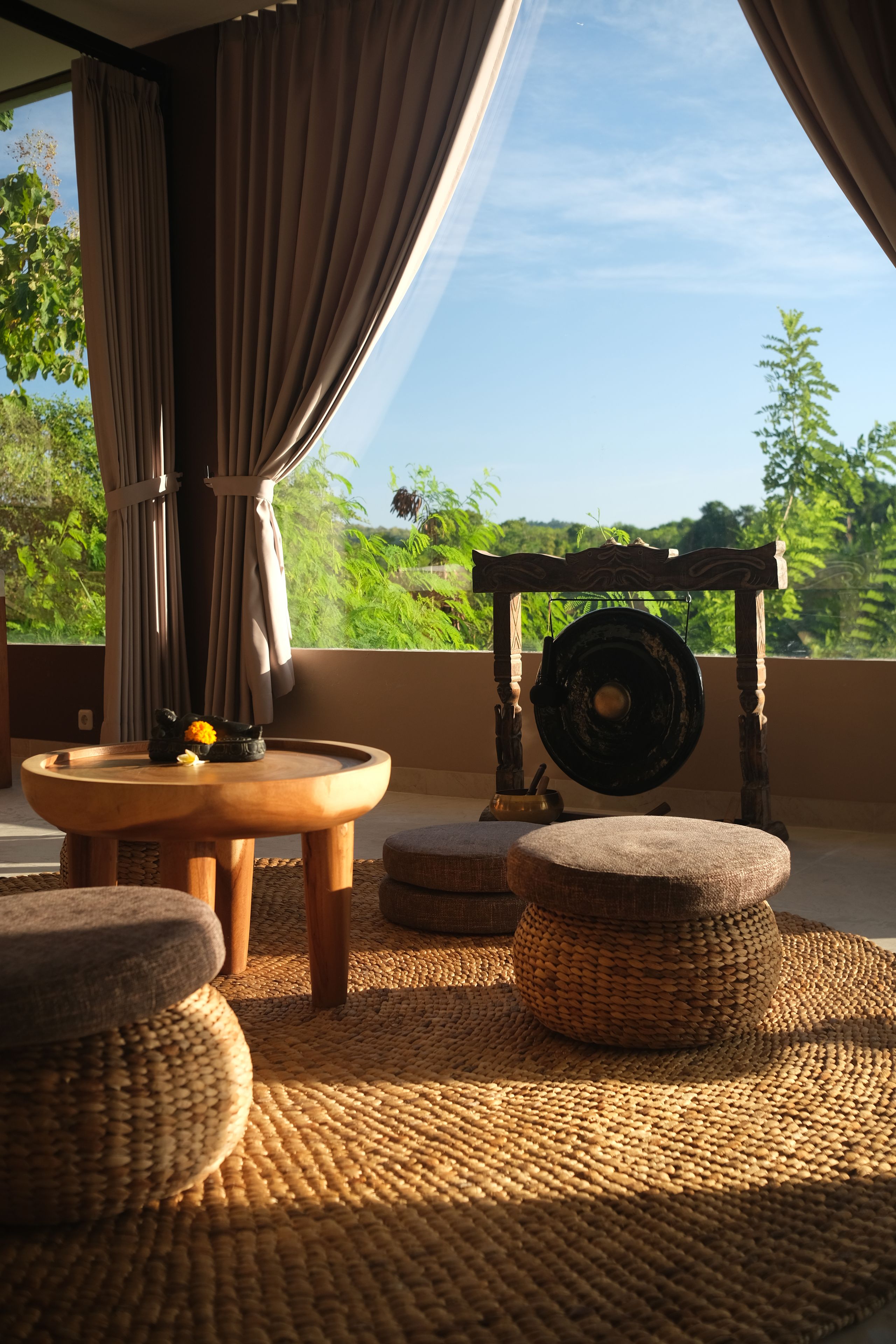 Quarto com vista para vegetação exuberante, uma mesa de madeira redonda com uma tigela e uma flor, e um tear tradicional.