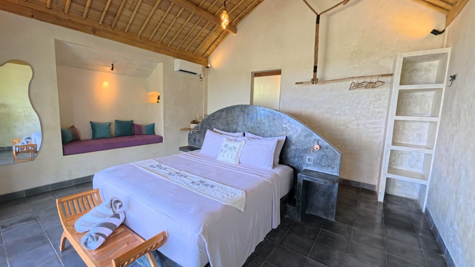 Quarto com cama, mesa de cabeceira, banquinho e prateleira na parede.