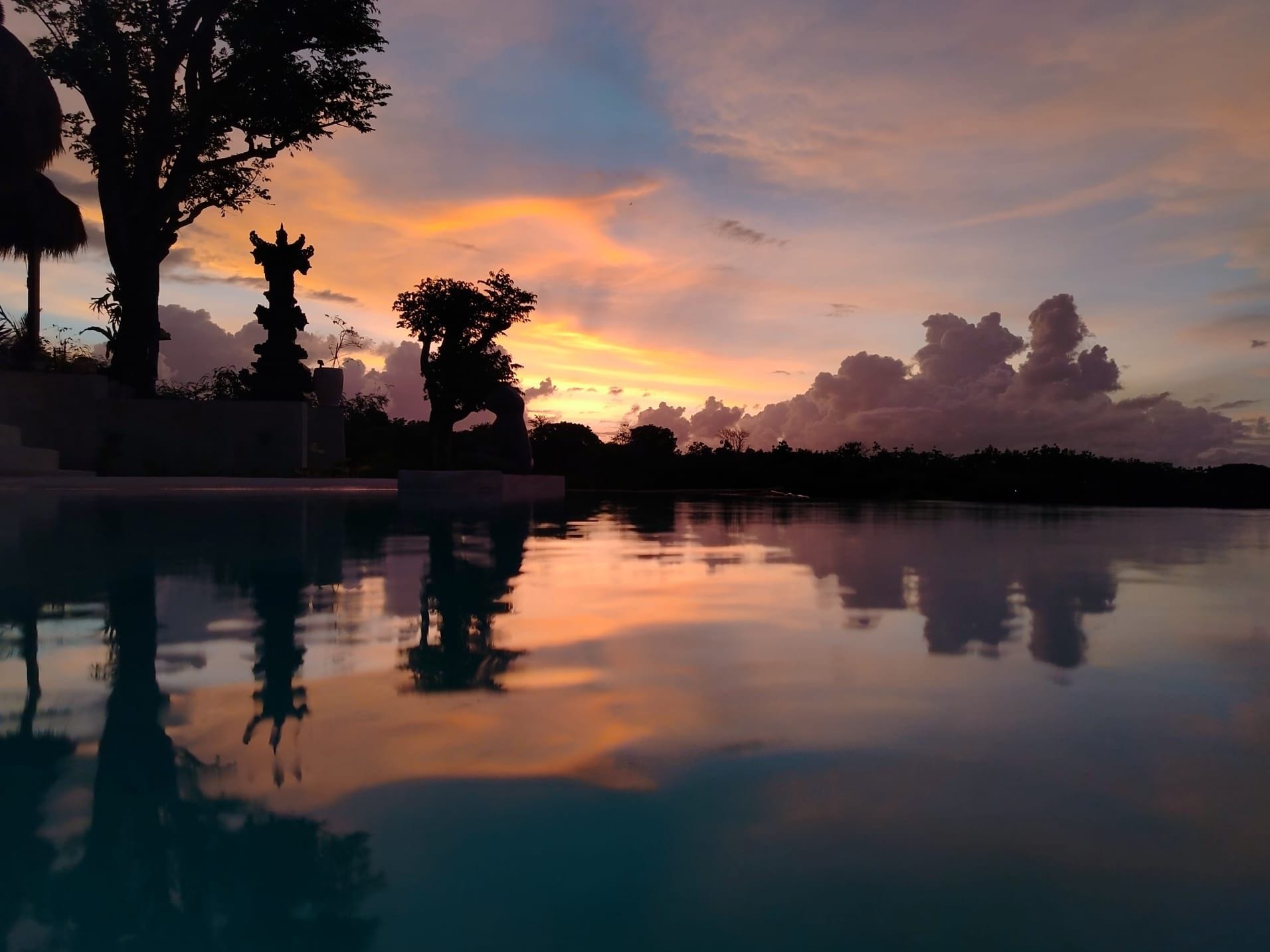 Um pôr do sol tranquilo sobre águas calmas, com reflexo, cercado por árvores e uma estátua.