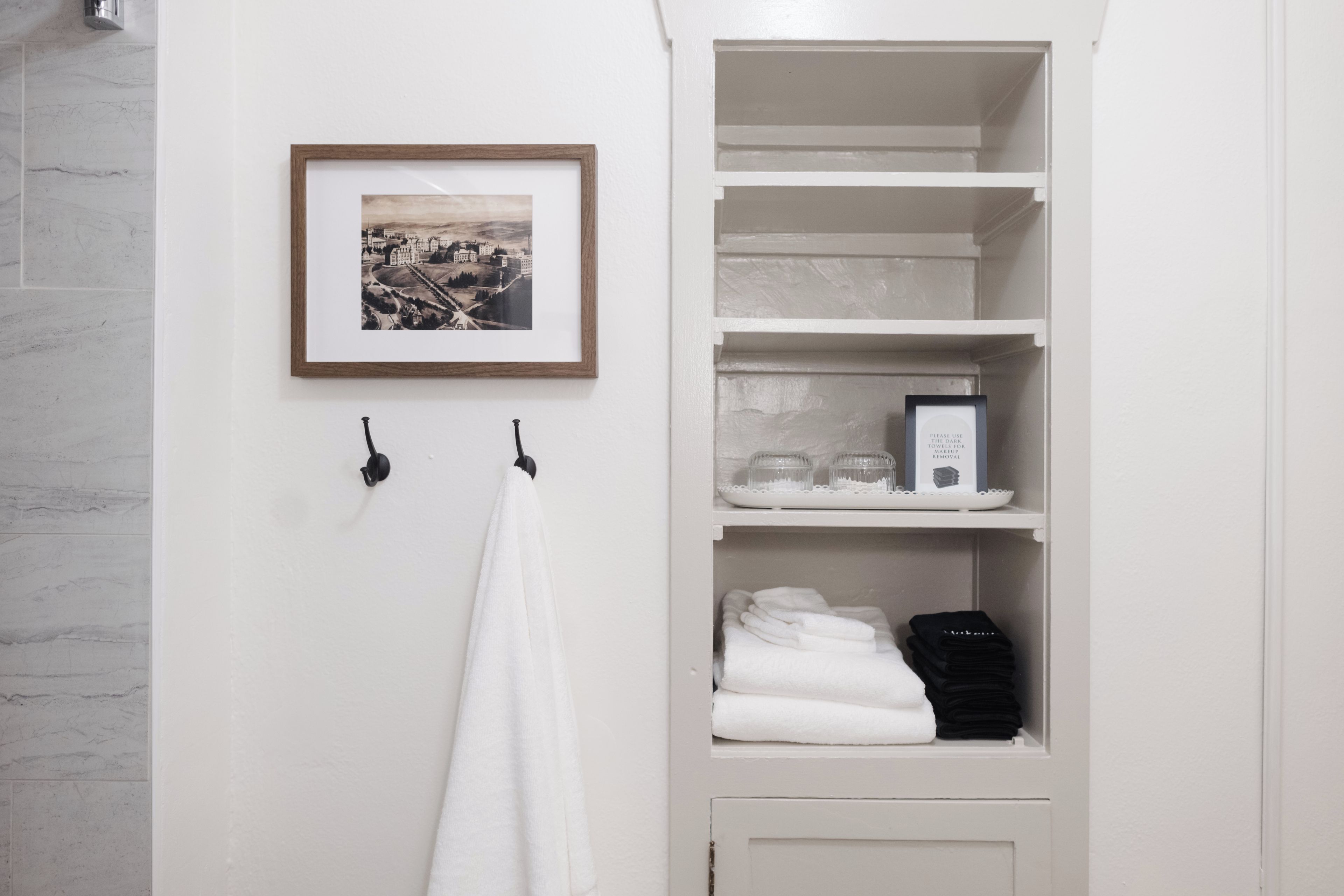 Ein Badezimmer mit einem weißen Handtuch an einem Haken, einem gerahmten Bild an der Wand und einem Ablagefach mit Handtüchern und einem Schild zur Entfernung von Make-up.