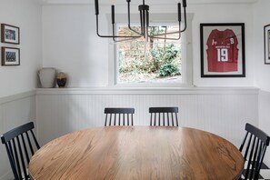 Een eetruimte met een houten tafel, zwarte stoelen, een raam met uitzicht op groen en ingelijste sporttenue aan de muur.