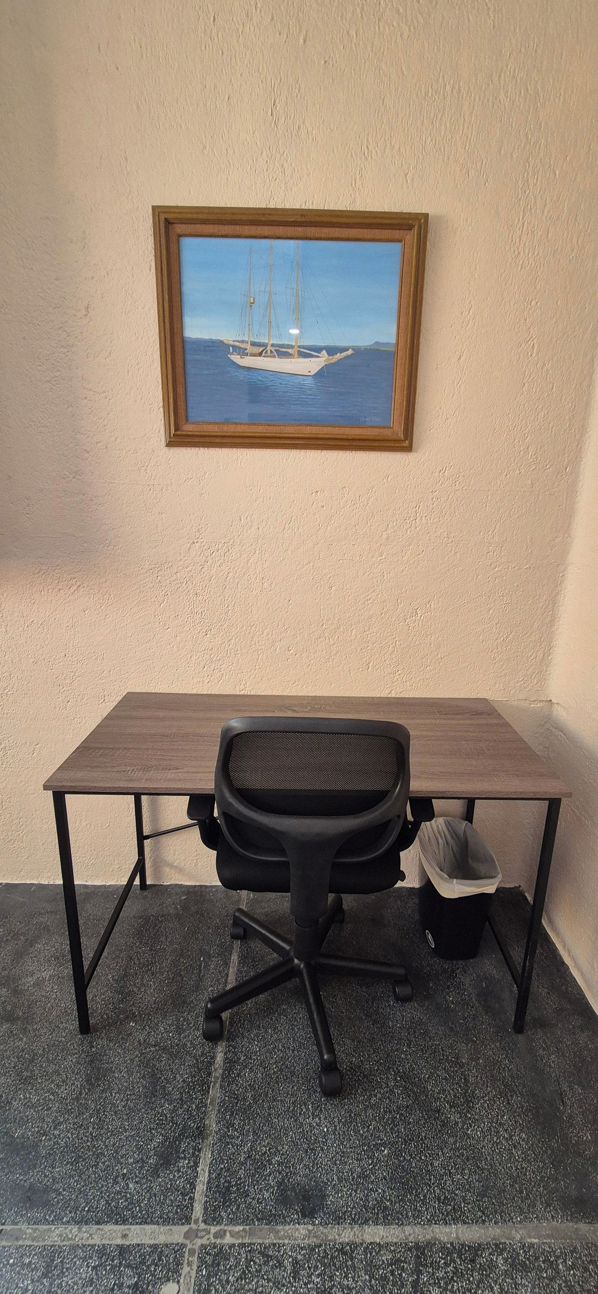 A desk with a chair and a trash bin, a framed painting of a sailboat on the wall, and a textured floor.