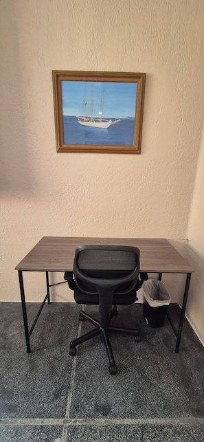 Et skrivebord med en stol og en skraldespand, et billede i ramme af en sejlbåd på væggen, og et struktureret gulv.