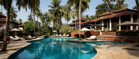 Una piscina con sillones de descanso y sombrillas, rodeada de palmeras y un edificio con tejado de baldosas.