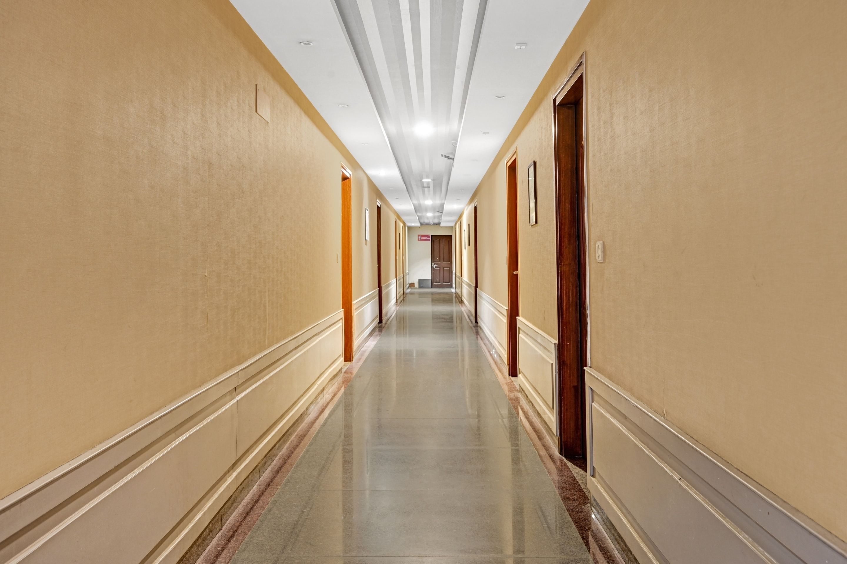 A long, narrow hotel corridor with doors on both sides, a shiny tiled floor, and a ceiling with recessed lighting.