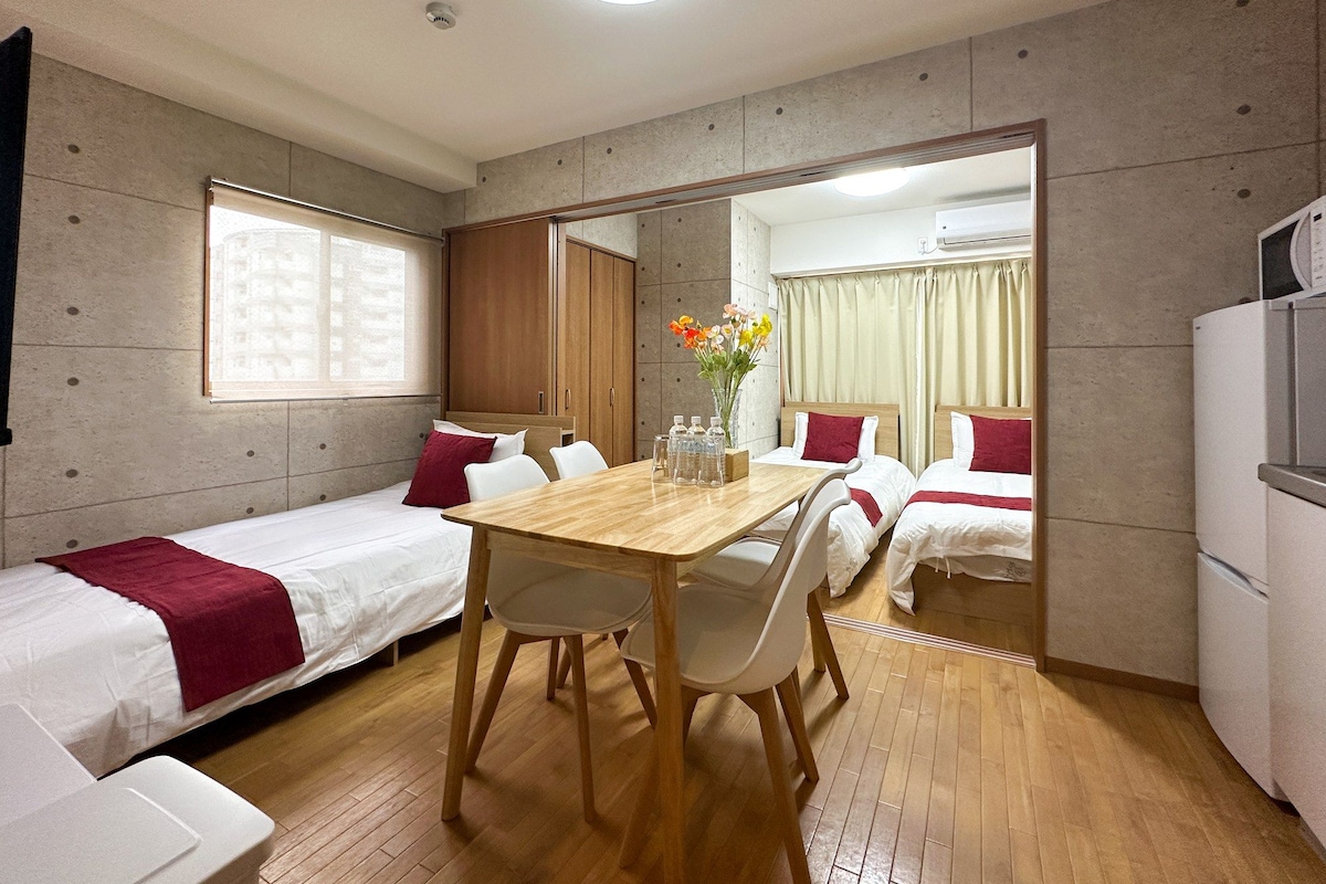 A compact hotel room with a wooden dining table, two single beds, a large mirror, and a window with blinds.
