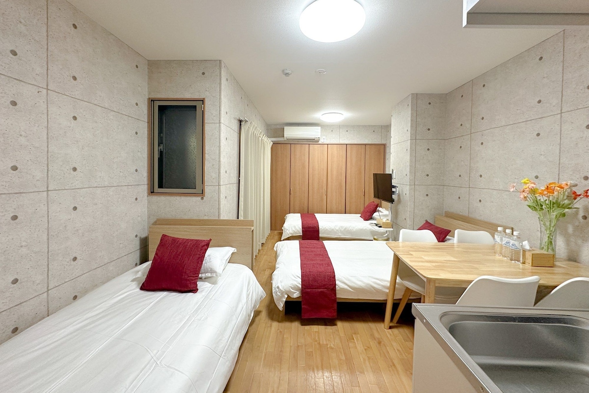 A hotel room with two beds, a wooden desk, a small kitchen area, and a bathroom visible in the background.