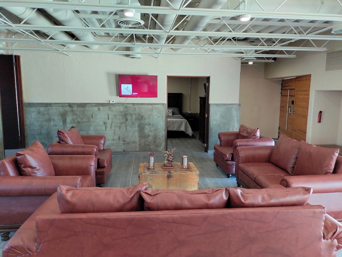 Un vestíbulo de hotel con sofás de cuero dispuestos alrededor de una mesa de centro, un televisor montado en la pared y una puerta de madera que conduce a otra habitación.