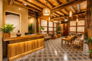 Un mostrador de recepción de madera con suelo de baldosas, una zona de estar con mesas y sillas, y un techo de madera con vigas a la vista.