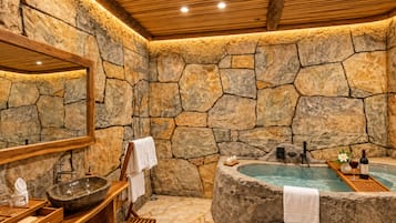 Un baño con paredes de piedra, una bañera independiente, un tocador de madera y un espejo grande.