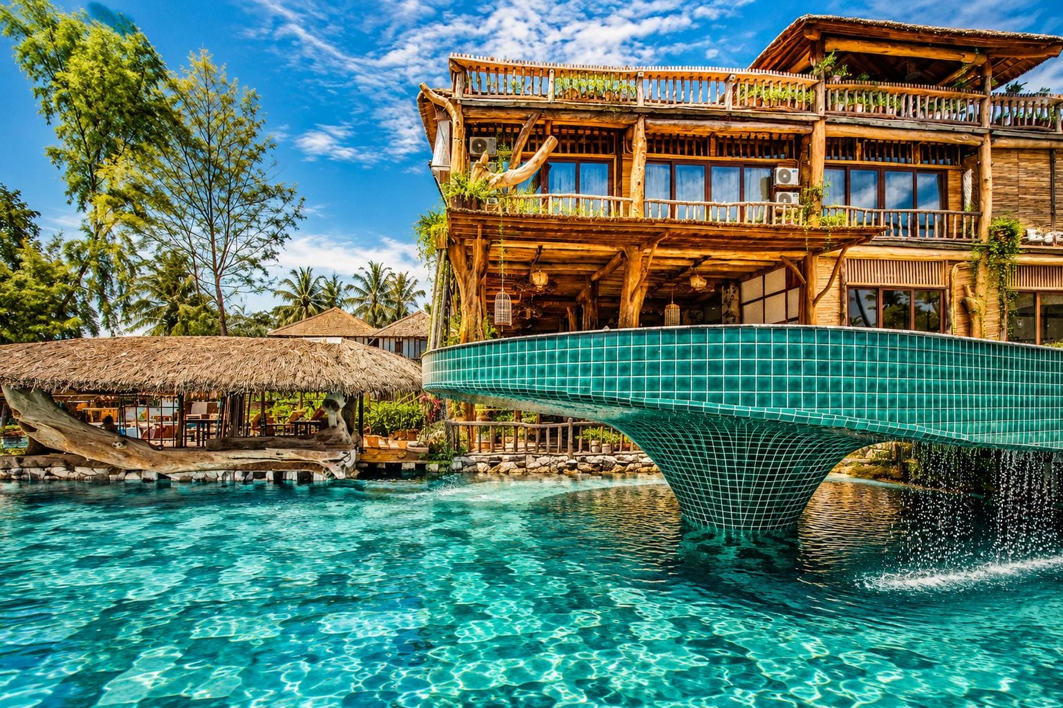 Ein Resort mit Schwimmbad, einer Holzbrücke und einem Gebäude mit Reetdach.