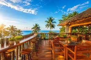 Una terraza de madera con zona de bar con vista a una playa, rodeada de palmeras y una estructura con techo de paja.