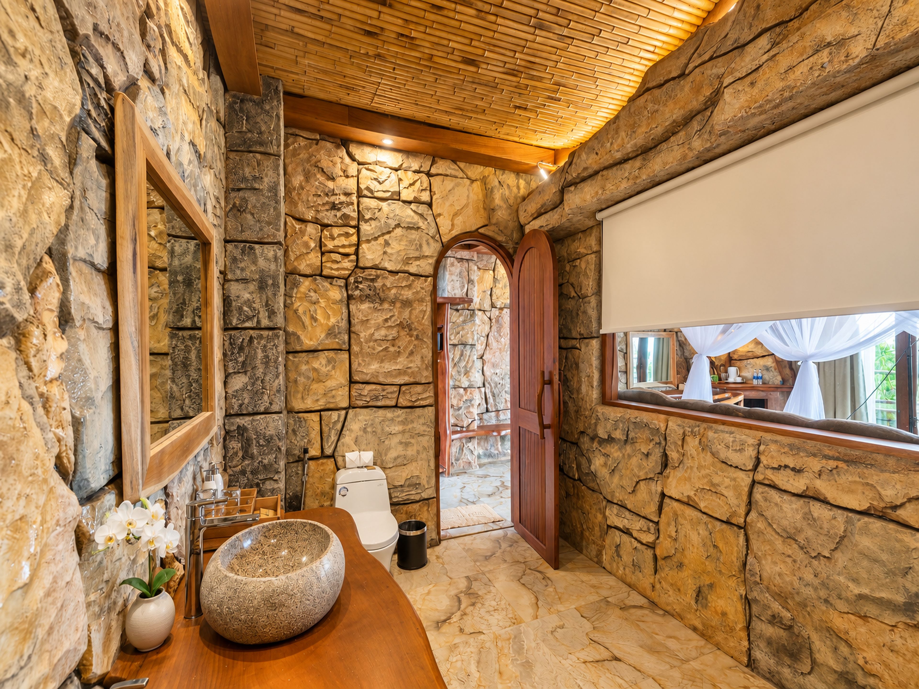 A bathroom with stone walls, a wooden ceiling, a large mirror, a sink with a stone basin, and a window with a view of the outside.