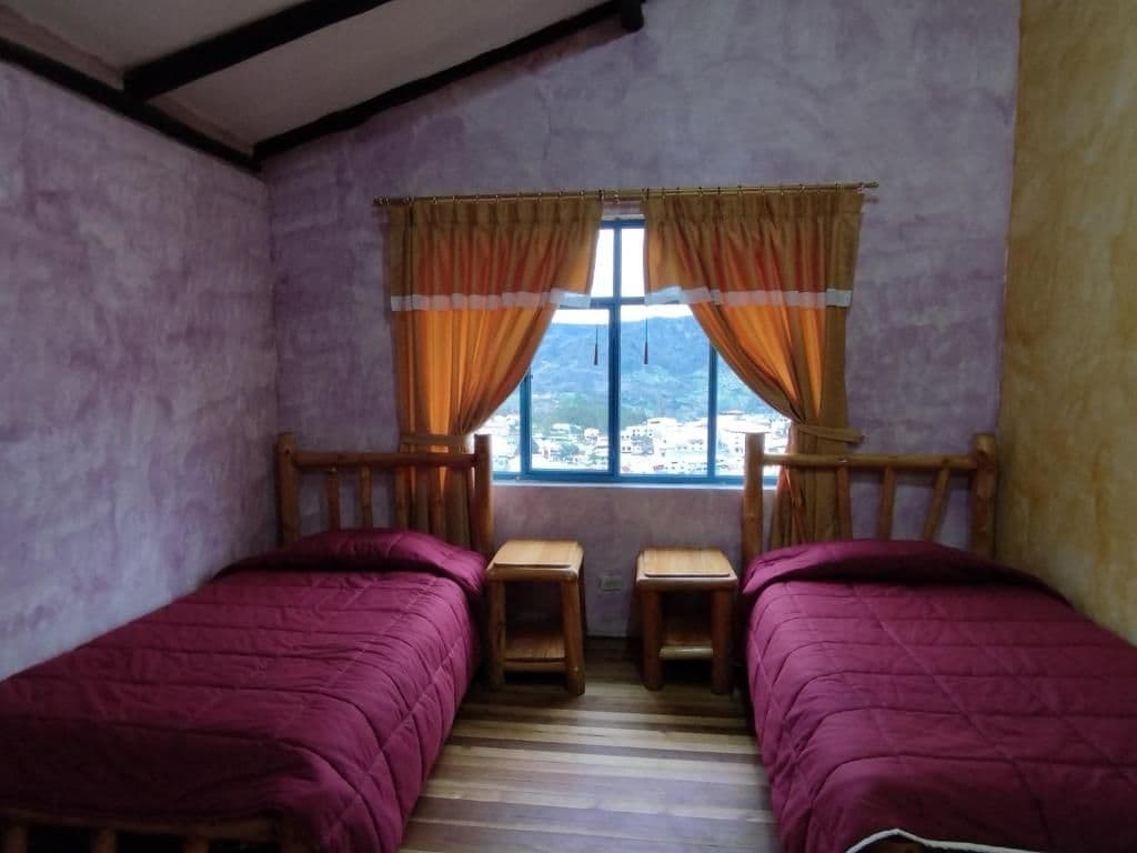 A room with two wooden beds, a window with curtains, and a view of a mountainous landscape.