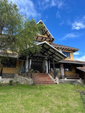 A two-story house with a steeply pitched roof, a covered porch, and a stone foundation. - Achik Wasi (Saraguro)