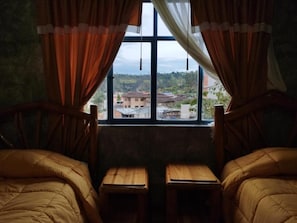 A room with two beds, a window with curtains, and a view of buildings outside.