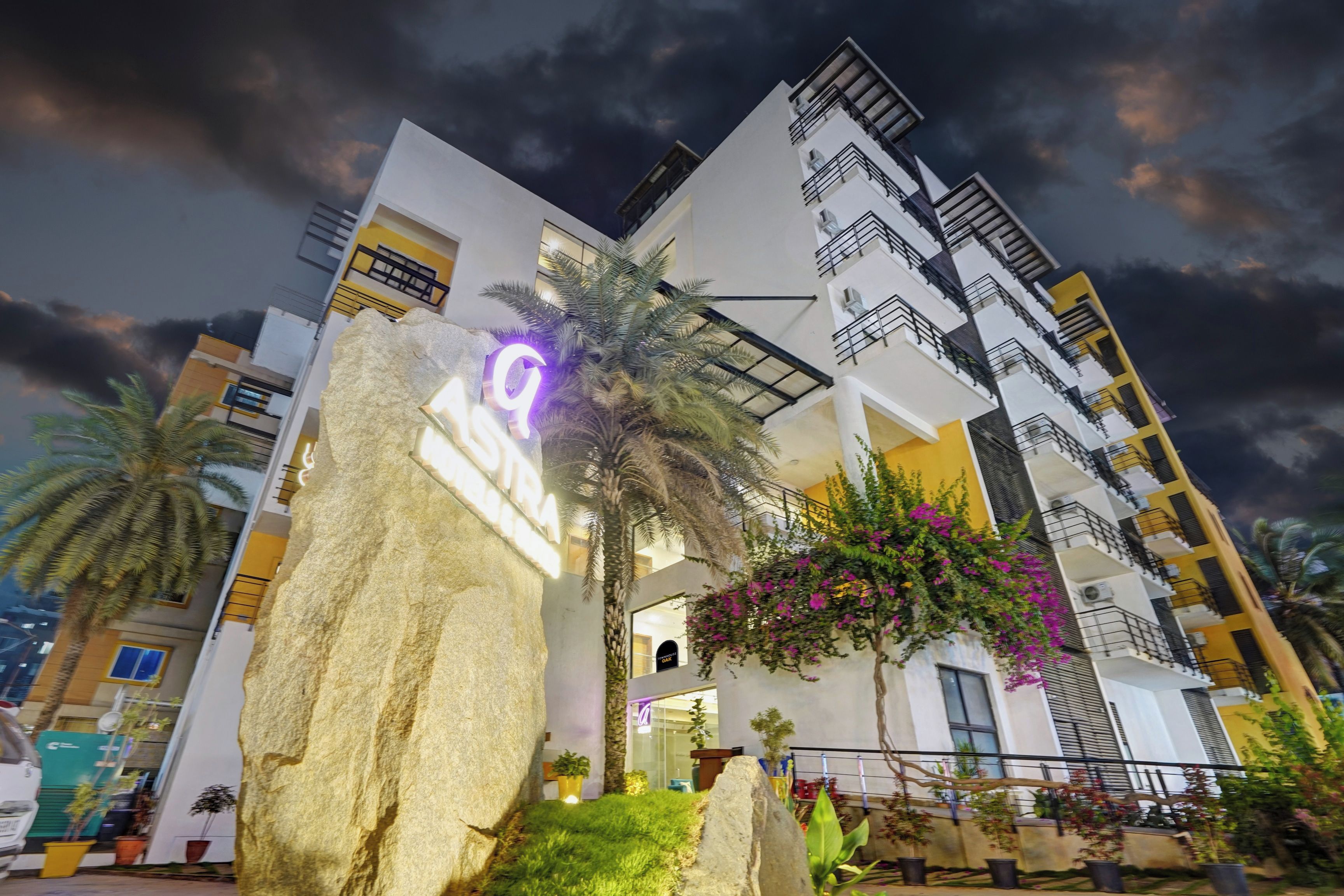 A multi-story hotel with a modern architectural design, featuring balconies and a well-maintained garden with palm trees and flowering plants.