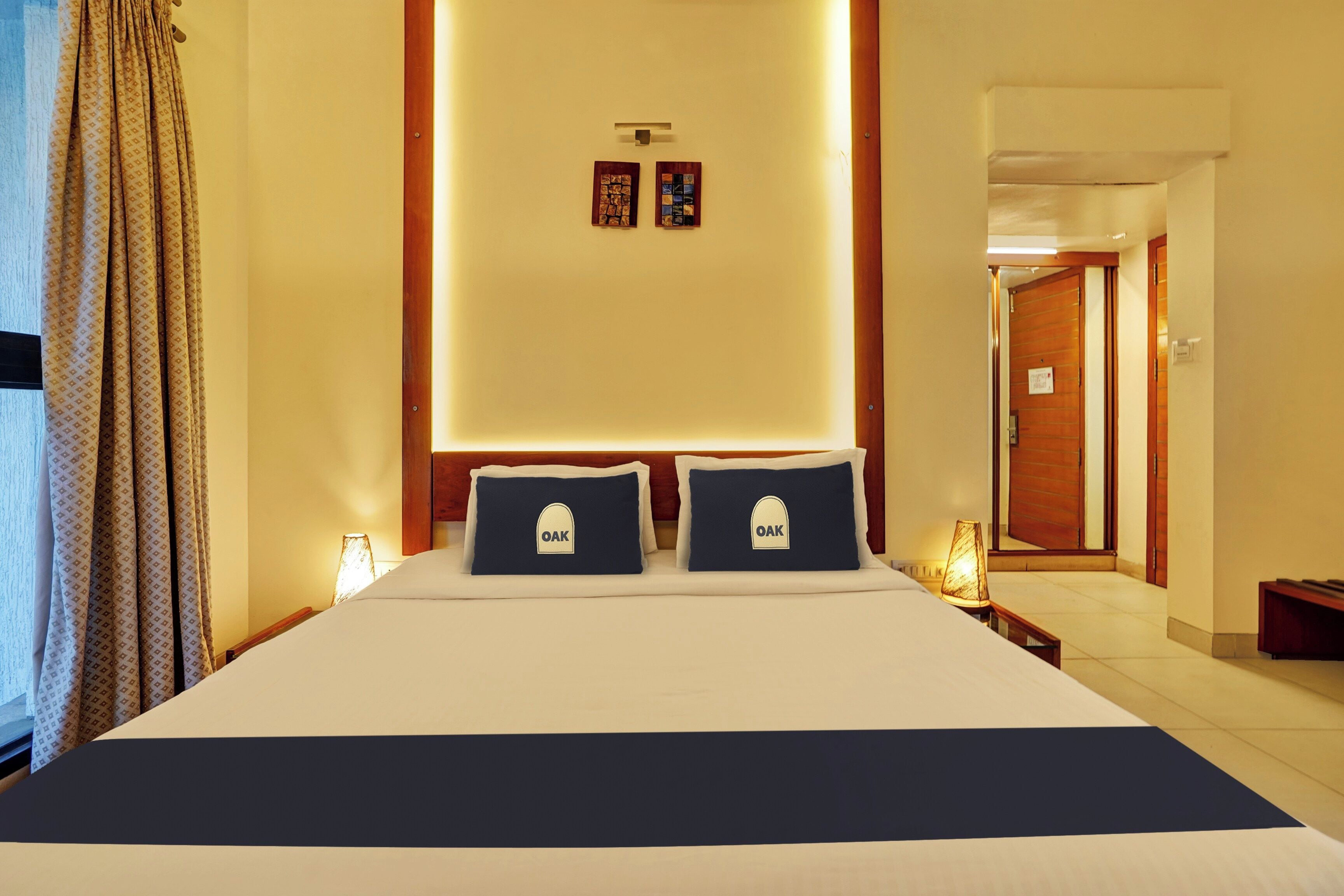 A hotel room with a large bed, two pillows with 'OAK' embroidered, a nightstand with a lamp, and a wall-mounted artwork.