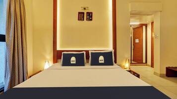 A hotel room with a large bed, two pillows with 'OAK' embroidered, a nightstand with a lamp, and a wall-mounted artwork.