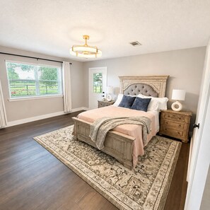 A bedroom with a large bed, a chandelier, a nightstand, and a view of the outdoors.