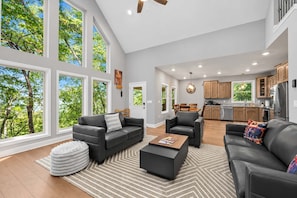 A spacious living area with a large window, a dining table, and a kitchen in the background. - Asheville Retreat | Hot Tub • Game Loft • Views! (Asheville)