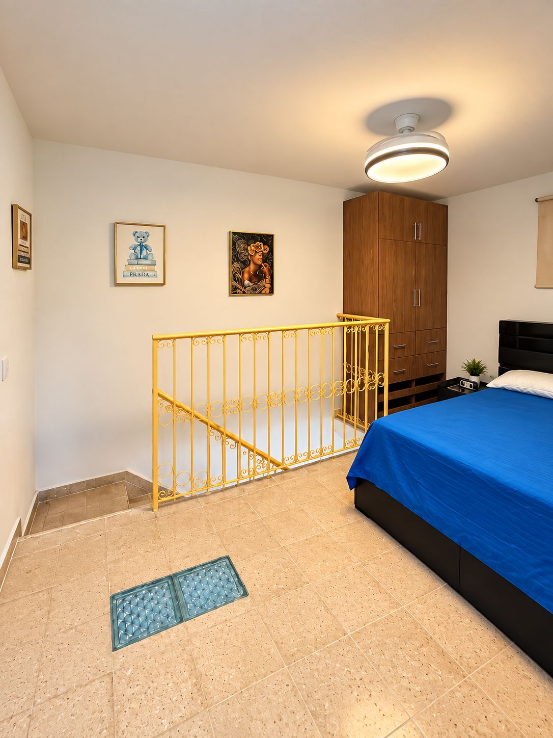 Un dormitorio con cama, armario, una mesita, un cuadro enmarco, barandilla decorativa y una lámpara en el techo.