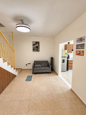 Ein modernes Interieur mit Treppe, einem grauen Sofa, Deckenventilator und einer durchgehenden Tür erkennbaren Küche.