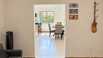 Un salon moderne avec un canapé gris, un coin repas comprenant une table et des chaises, et une cuisine visible à travers une porte ouverte.