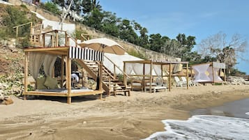 Overnatning ved stranden med trækonstruktioner, herunder seng og liggestole, foran en bakke og træer.