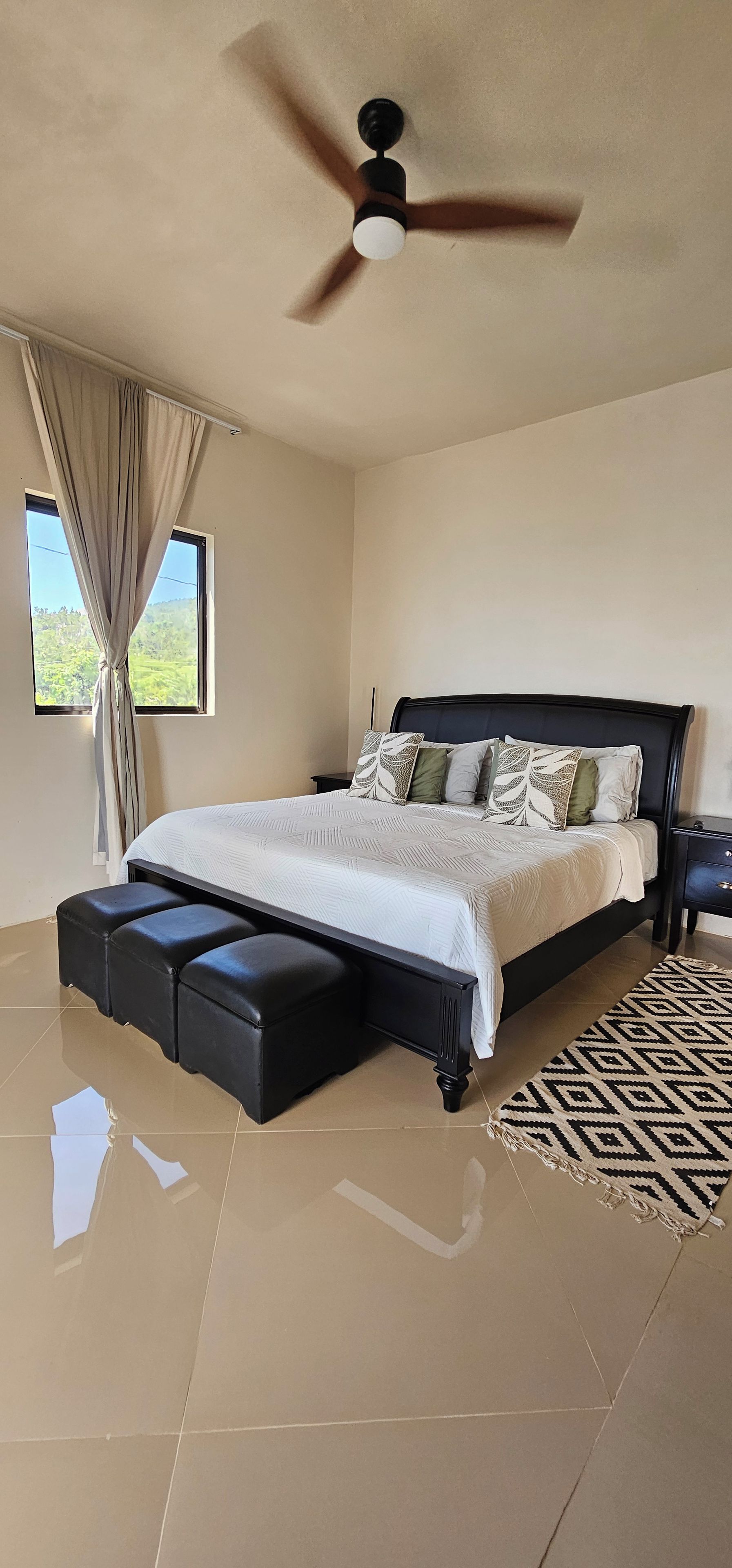 Une chambre à coucher avec un lit, un ventilateur de plafond, une fenêtre avec des rideaux et un tapis à motifs.