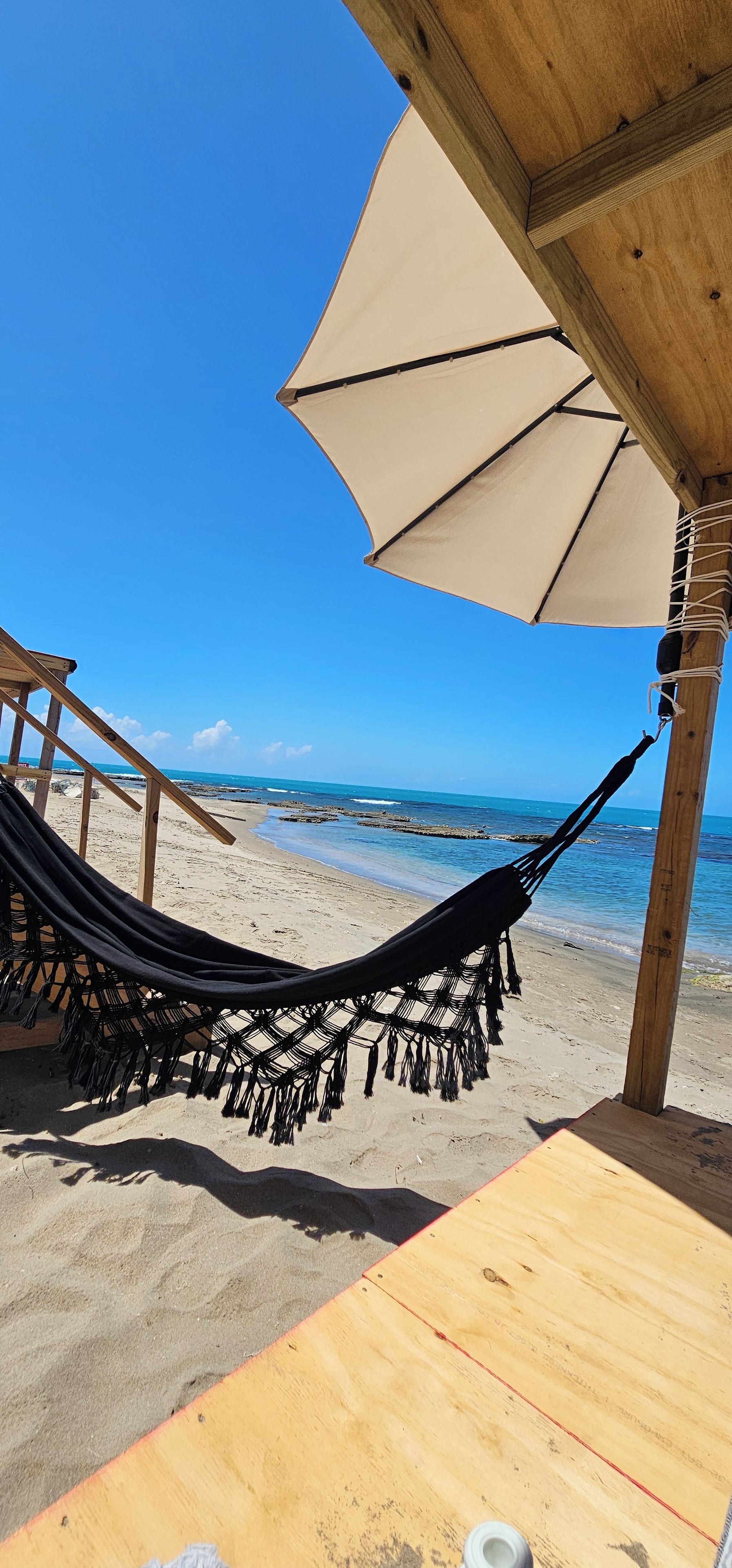 Une scène de plage avec un hamac sous une structure en bois, une table et une vue sur l’océan.