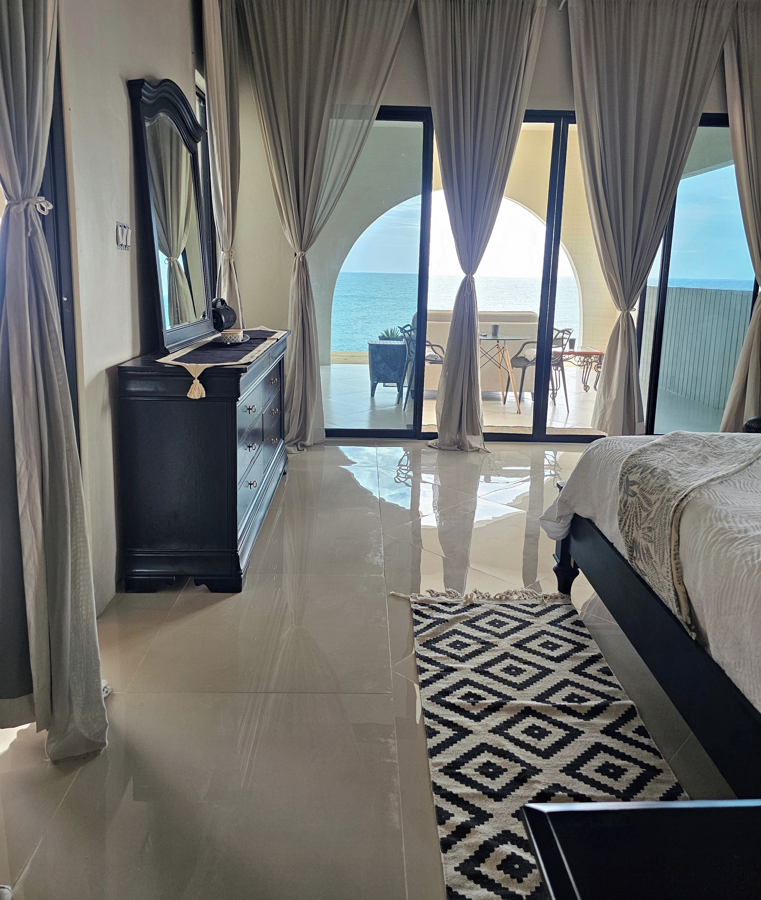 A bedroom with a bed, a dresser, a mirror, and a view of the sea through large windows.