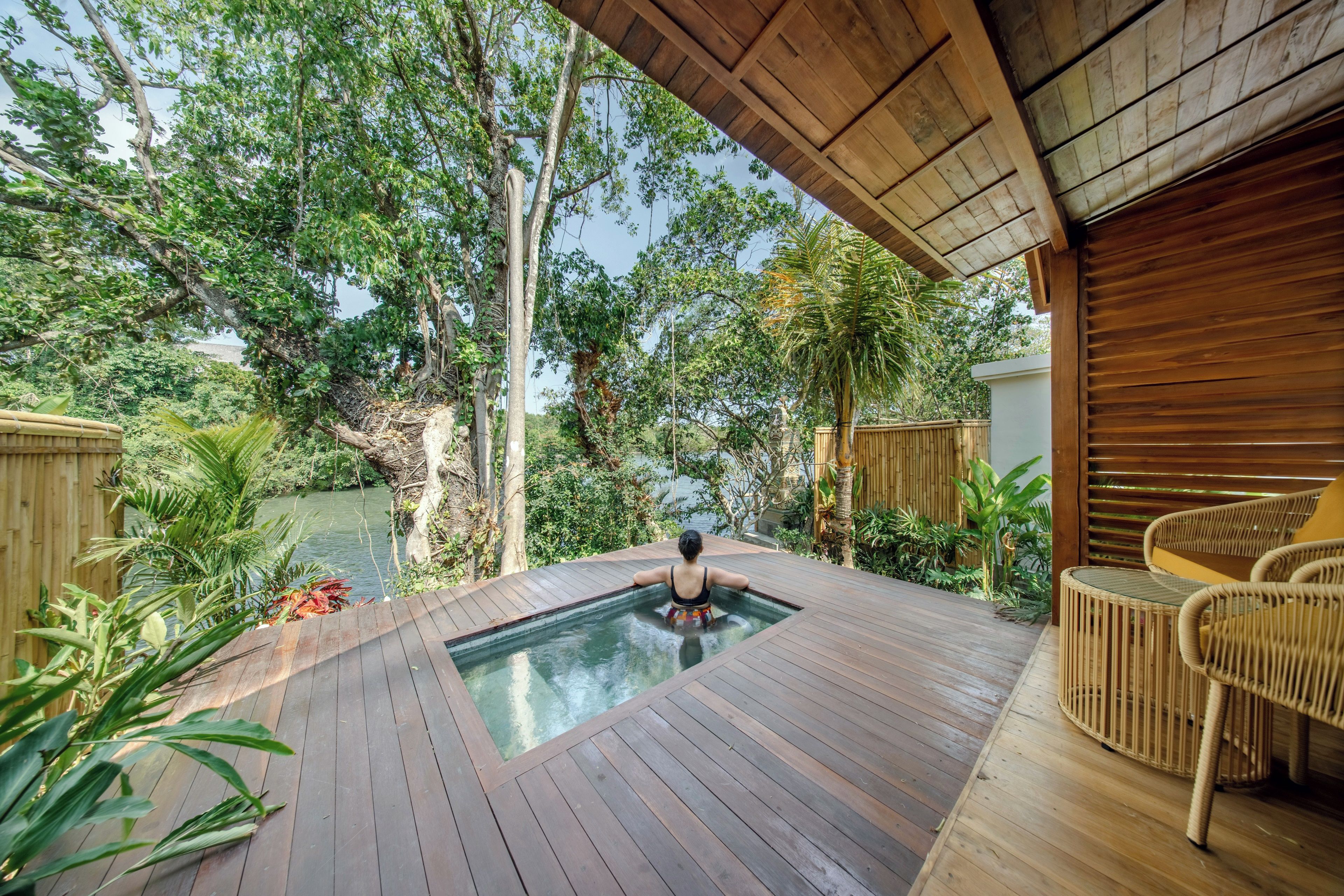 A wooden deck with a hot tub, surrounded by lush greenery and a view of a river.