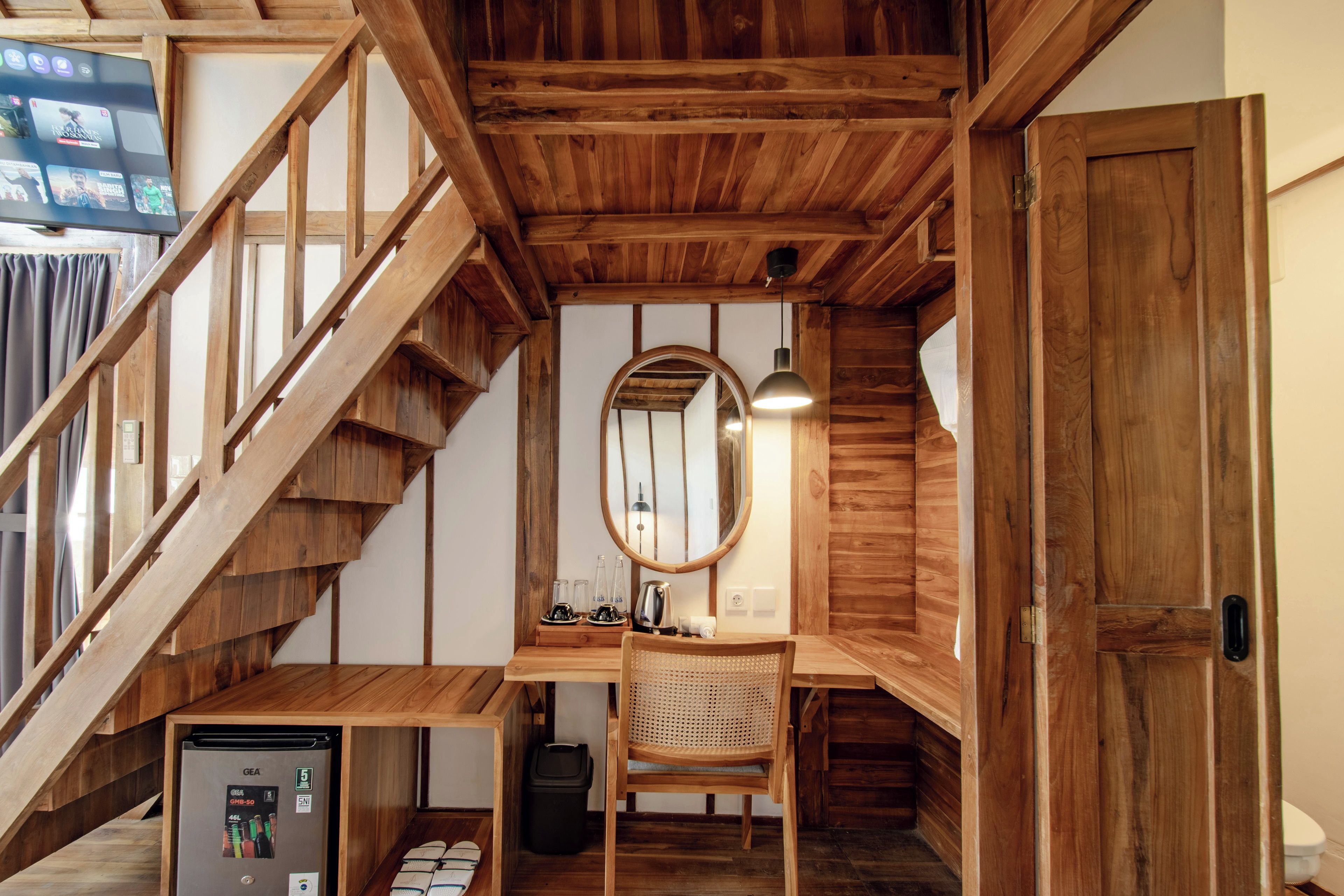 A wooden staircase leading to a loft bedroom with a desk, chair, and a small refrigerator.