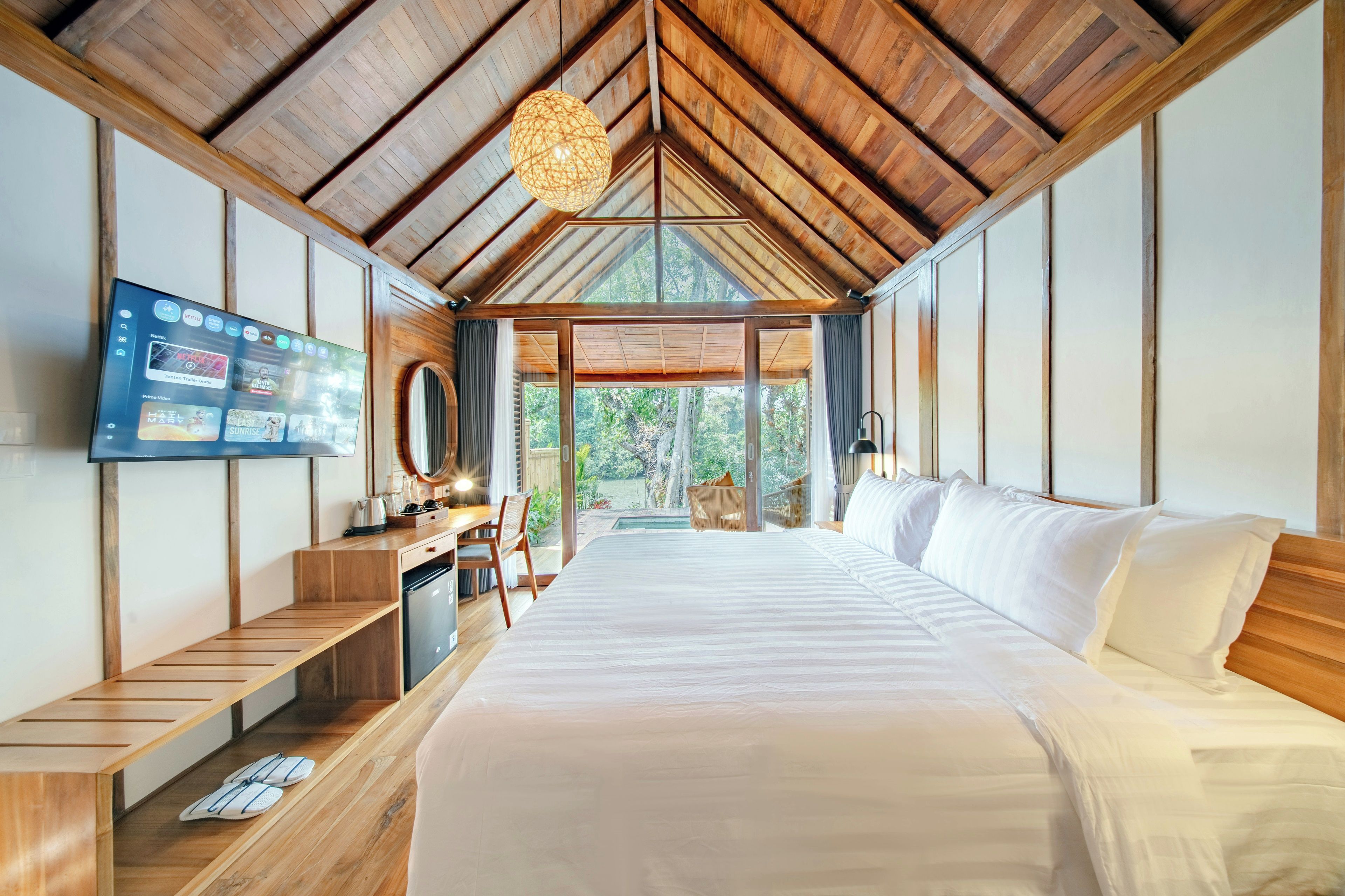 A bedroom with a large bed, a desk, a television, and a view of the outdoors.