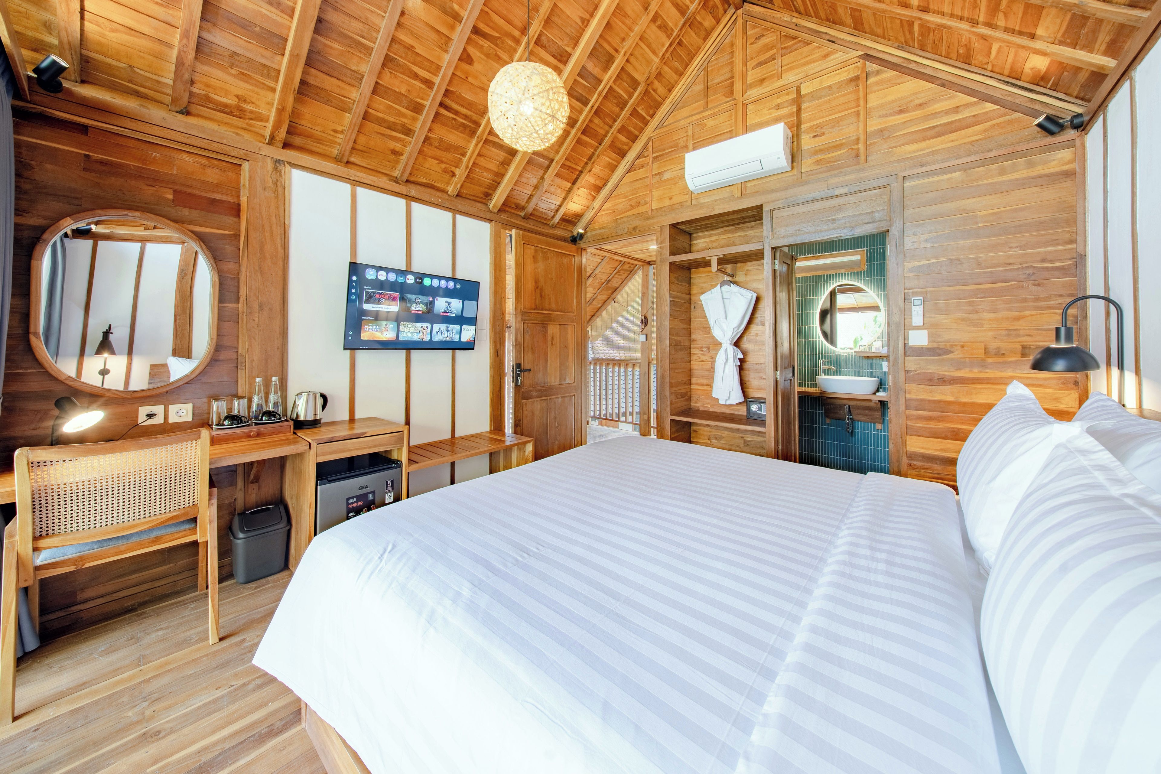 Un dormitorio tipo cabaña de madera con cama, escritorio, televisor y baño visible a través de una puerta abierta.