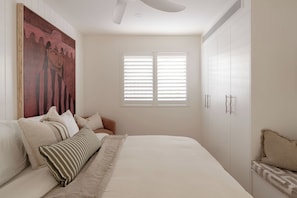 Une chambre à coucher avec un lit, des oreillers, un grand tableau, une fenêtre avec des stores et une armoire.