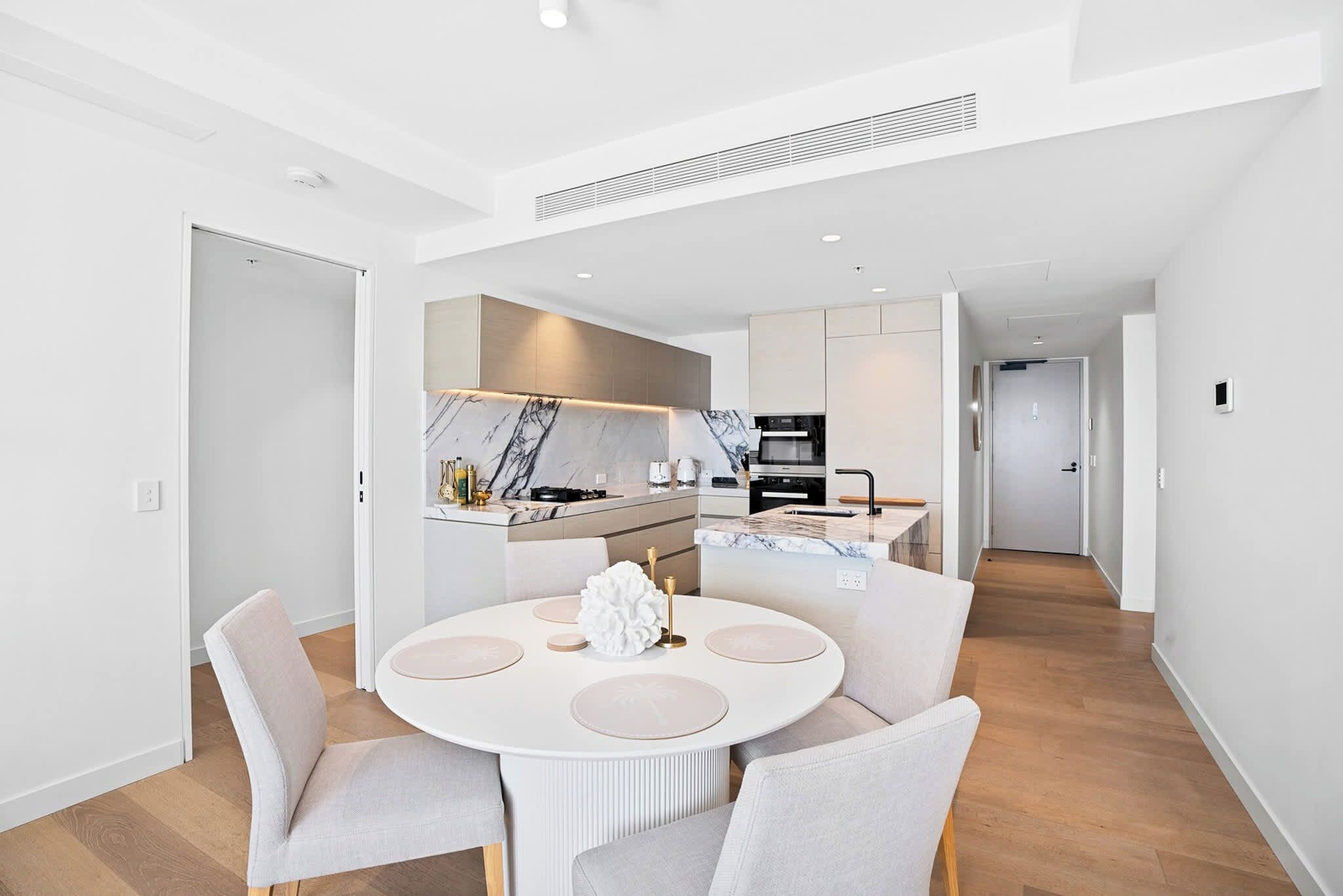 A modern kitchen with a central island, a dining area with a round table and chairs, and a hallway leading to another room.