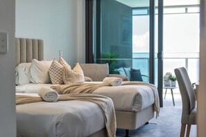 A hotel bedroom with a large bed, multiple pillows, a side table, and a view of the outdoors. - Jewel Designer Residence | Direct Ocean Views (Surfers Paradise)