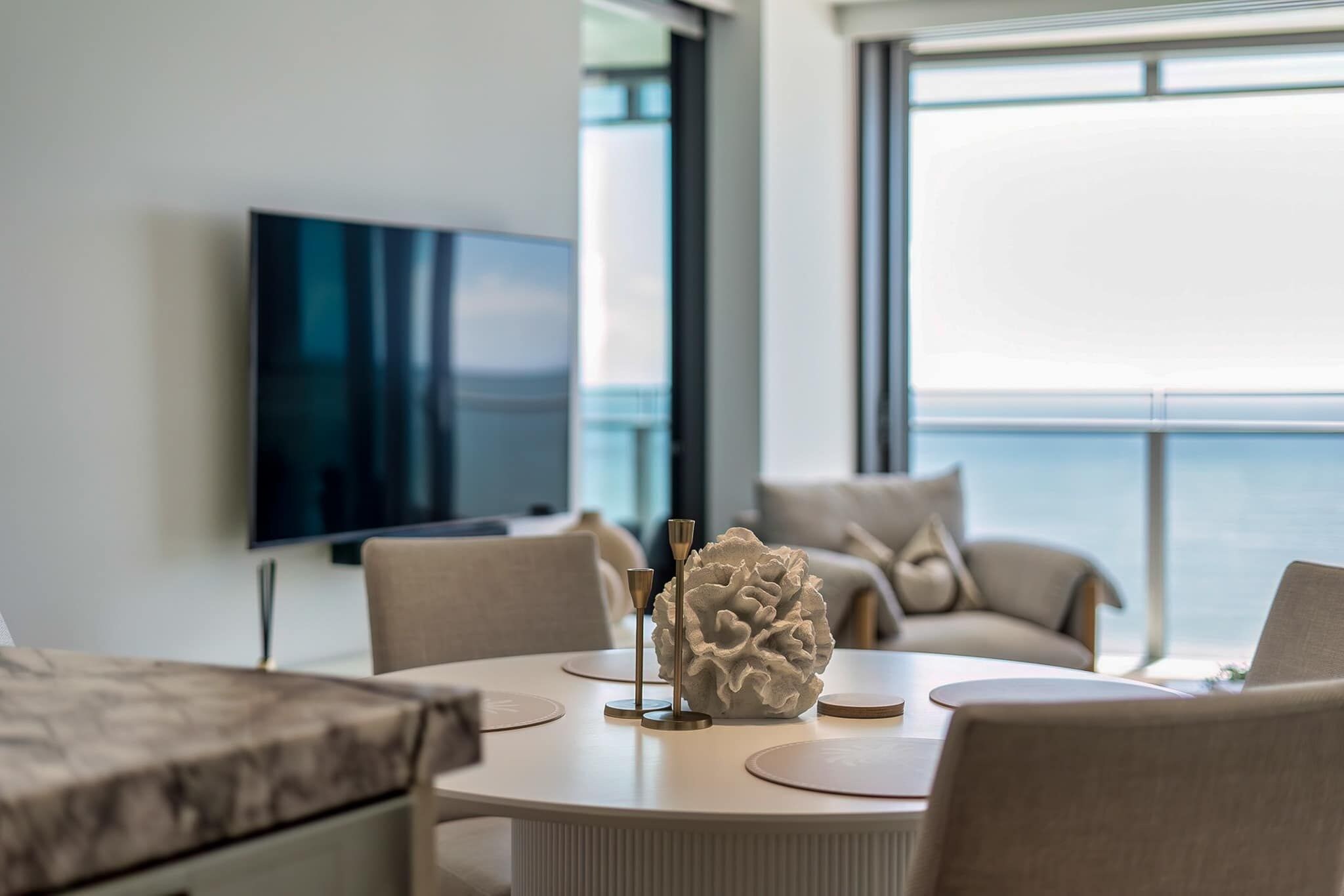 A modern living room with a round table, comfortable seating, a decorative centerpiece, and a large window offering a view of the sea.