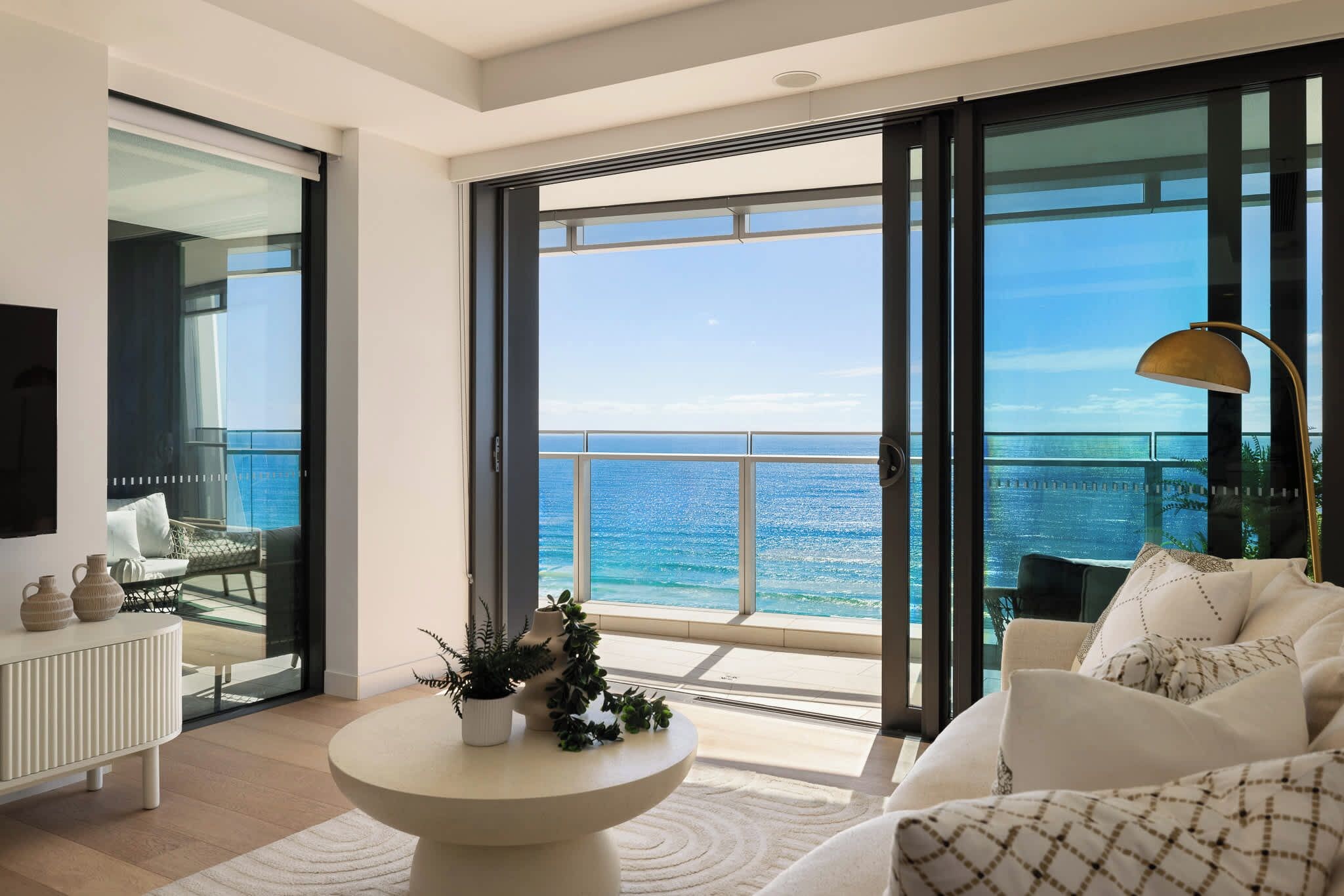 Een moderne woonkamer met een groot balkon met uitzicht op zee, een ronde salontafel met planten en een bank met gestreepte kussens.