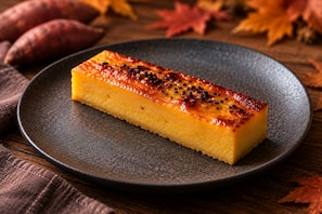 Un postre rectangular sobre un plato oscuro con superficie texturizada, rodeado de hojas otoñales y boniatos.