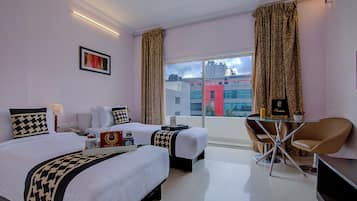 Habitación de hotel con dos camas, una zona de estar con una silla, una mesa y vistas a edificios a través del ventanal.