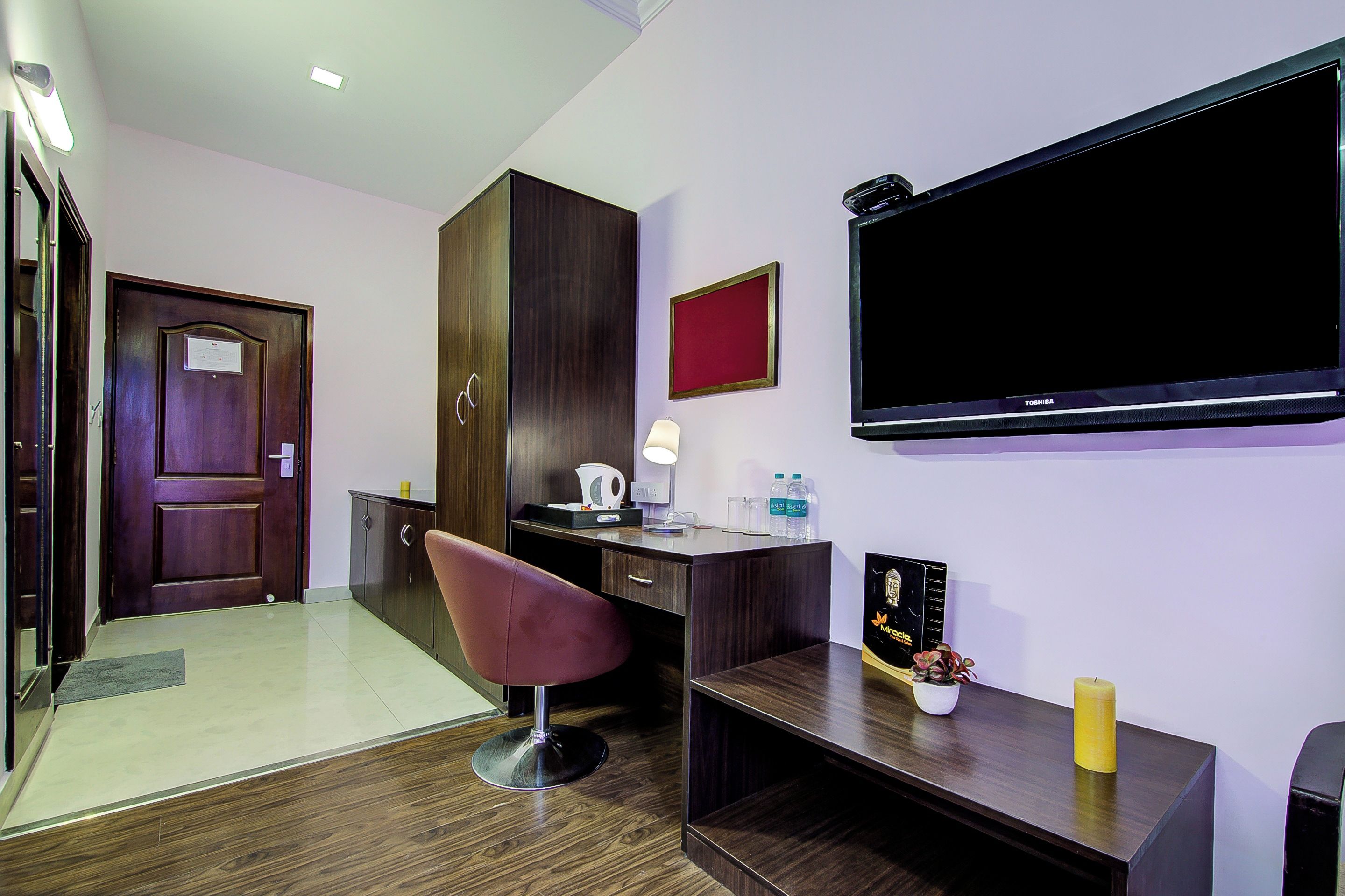 A hotel room with a wooden desk, a chair, a flat-screen TV, a red-framed picture, a small potted plant, and a candle on a shelf.