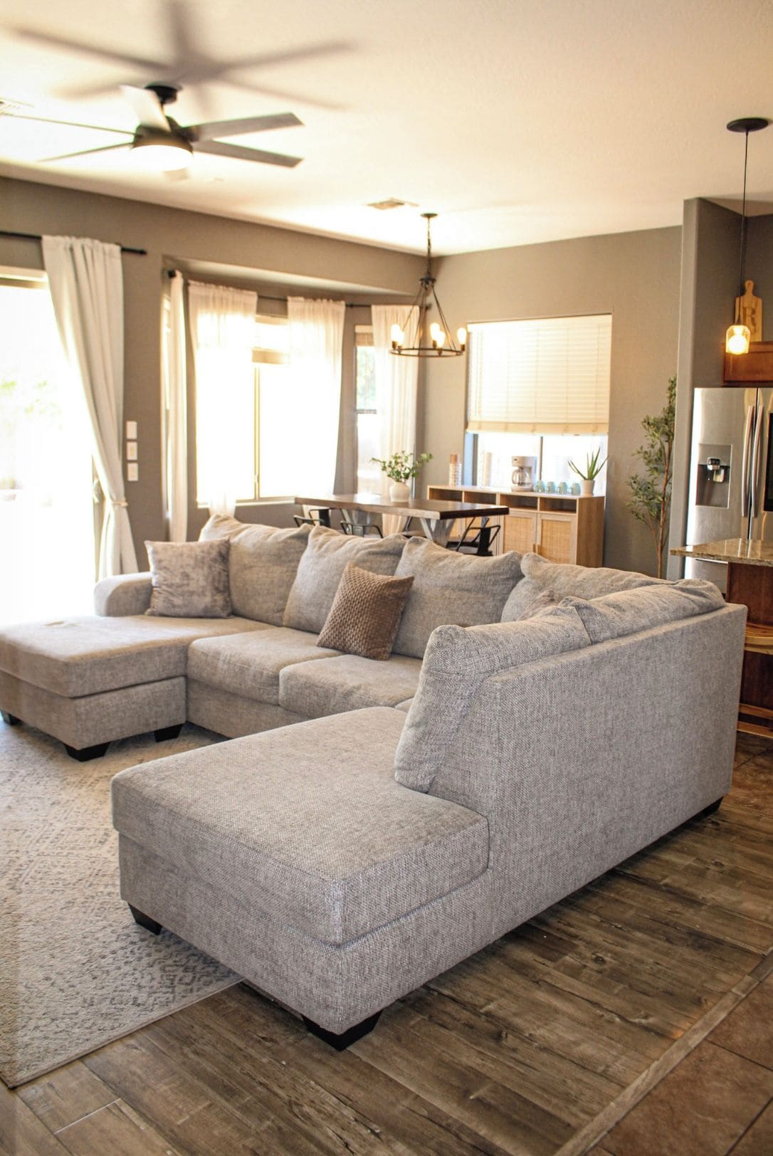 A modern living room with a sectional sofa, a ceiling fan, and a kitchen area in the background.